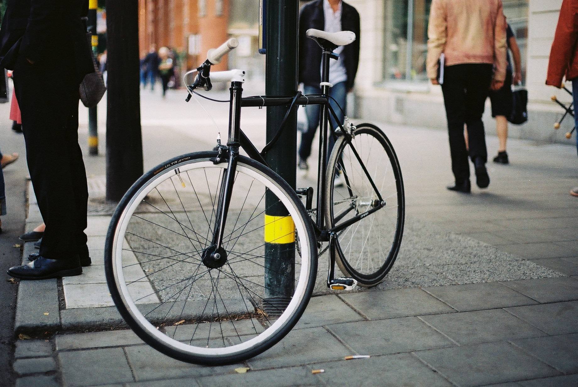 a blank and white single speed bicycle locked to the street post, several people walking on the sidewalk behind