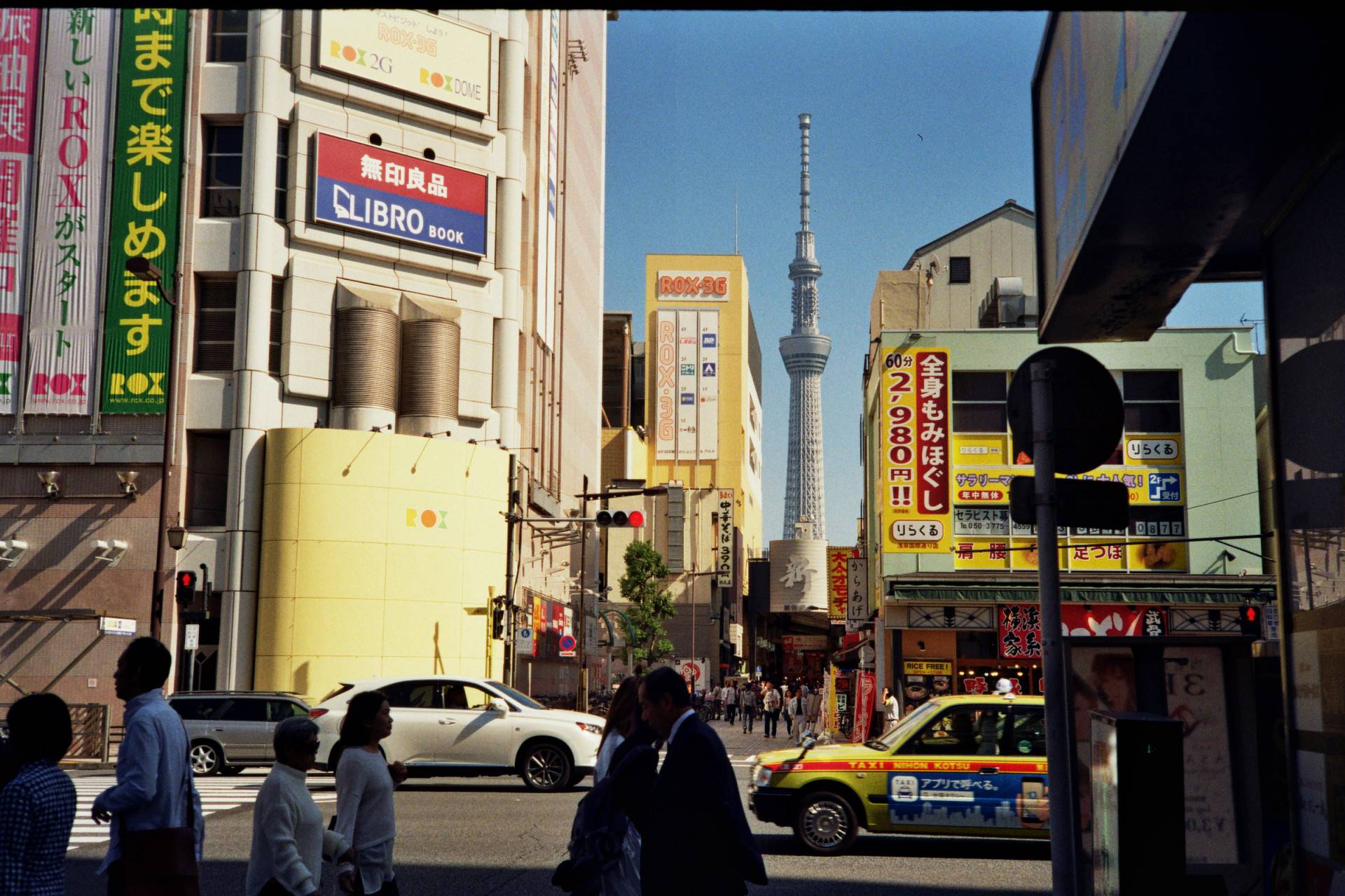 view of a busy pedestrian street seen from the other side of the intersecting main street, buildings around the street are covered with signs, there is a tower on the background