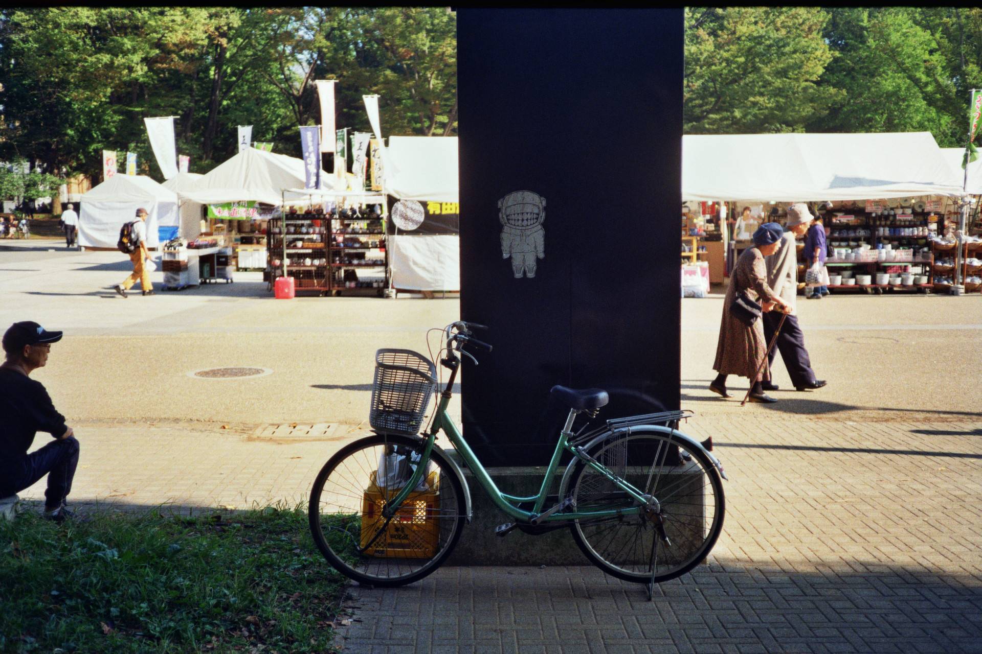 a bicycle resting in front of a dark pillar in the middle of the image, there is a small astronaut image on the pillar, a man is sitting on the left side looking at the market behind, an old couple is walking on the right side