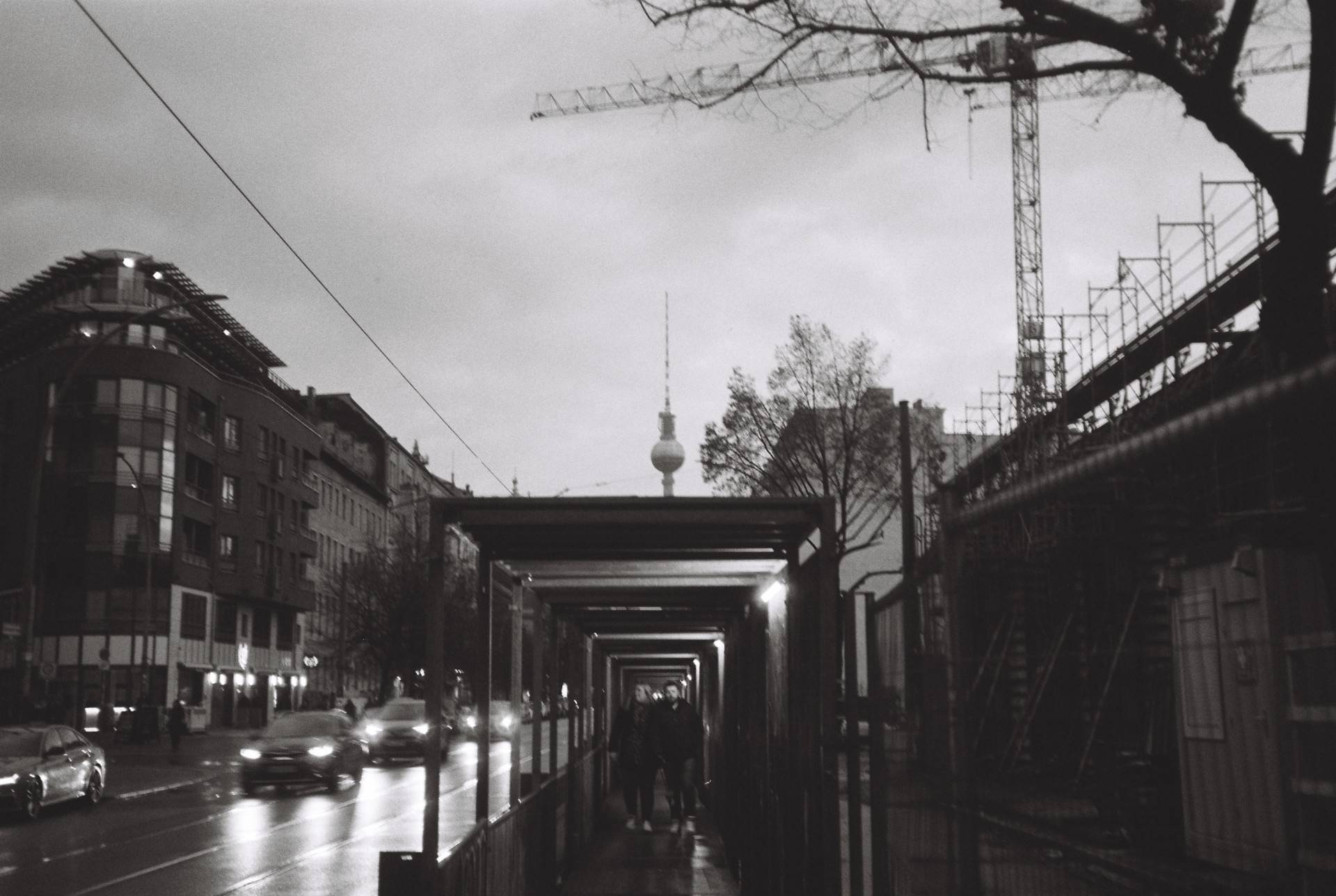 a couple walking towards the camera through a tunnel on the sidewalk due to the construction next to it, TV Tower is seen on the background under the grey cloudy sky