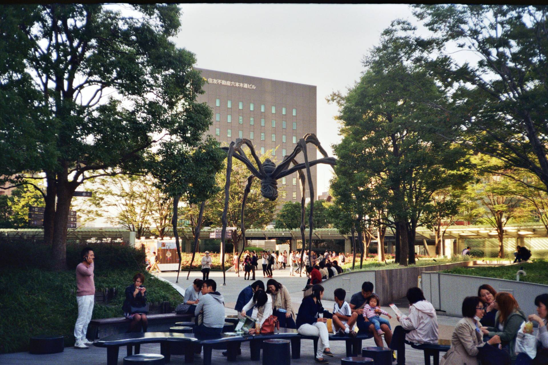 people hanging around in a park with trees on grass patches, there is large statue in the middle that resembles a spider, and a larger building on the background