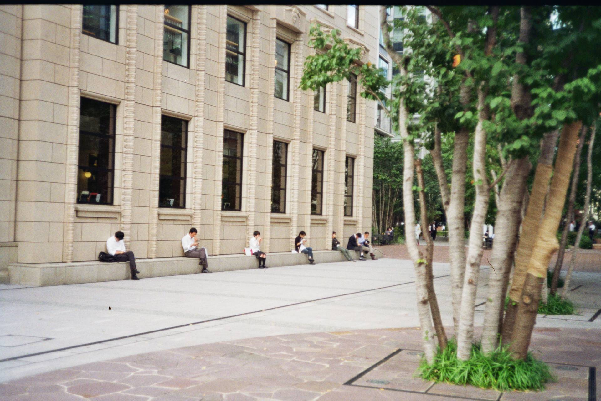 people sitting next to a building and eating their lunch, they sit almost equidistant from each other, a person below each window of the building