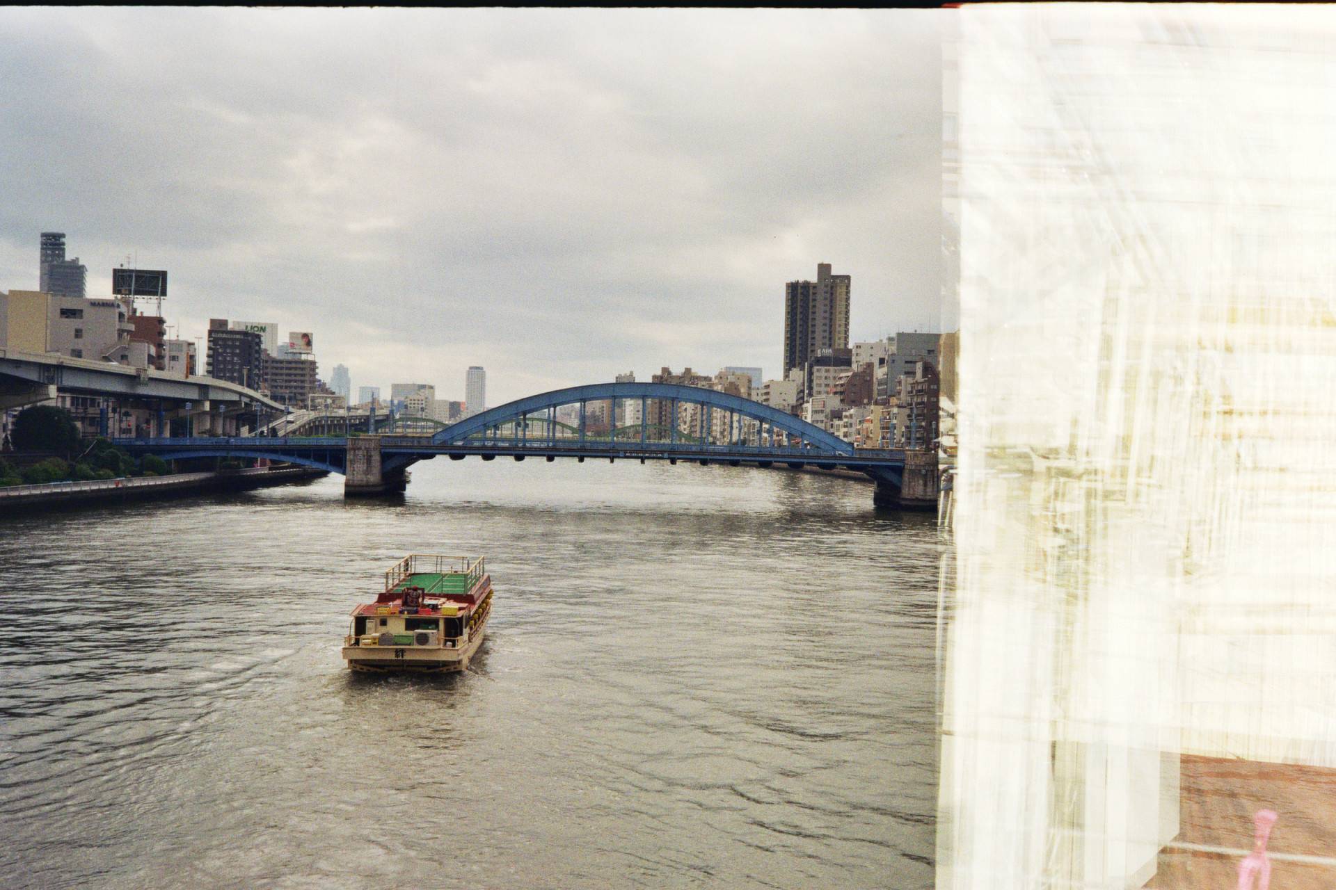 a small boat cruising along the river approaching the underpass below the bridge, river is surrounded with buildings, right side of the image has white artefacts from the burnt film