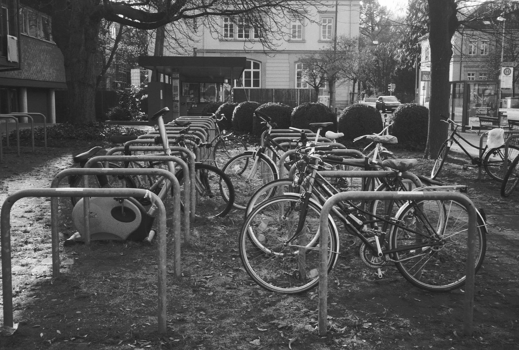 a parking area for bicycles covered with grass in front of a building, parking area is half full and one of the parked ones is an exercise bike