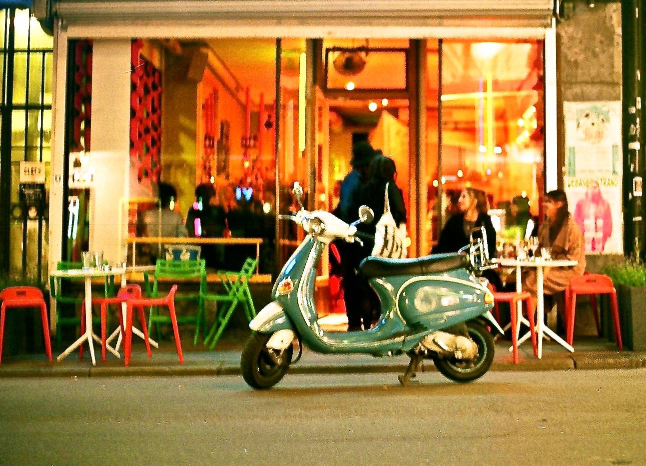 a turquoise vespa parked in front of a cafe where people are sitting outside 