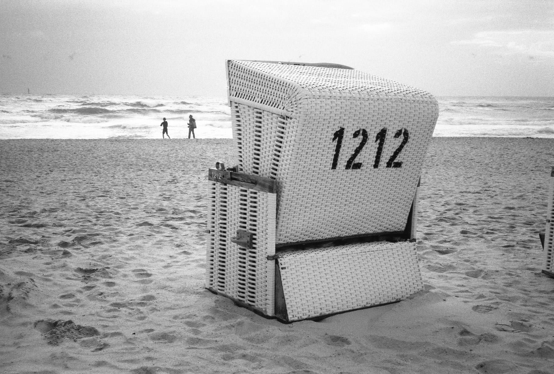 a beach basket chair numbered 1212 seen from behind, two people walking by the sea seen on the background