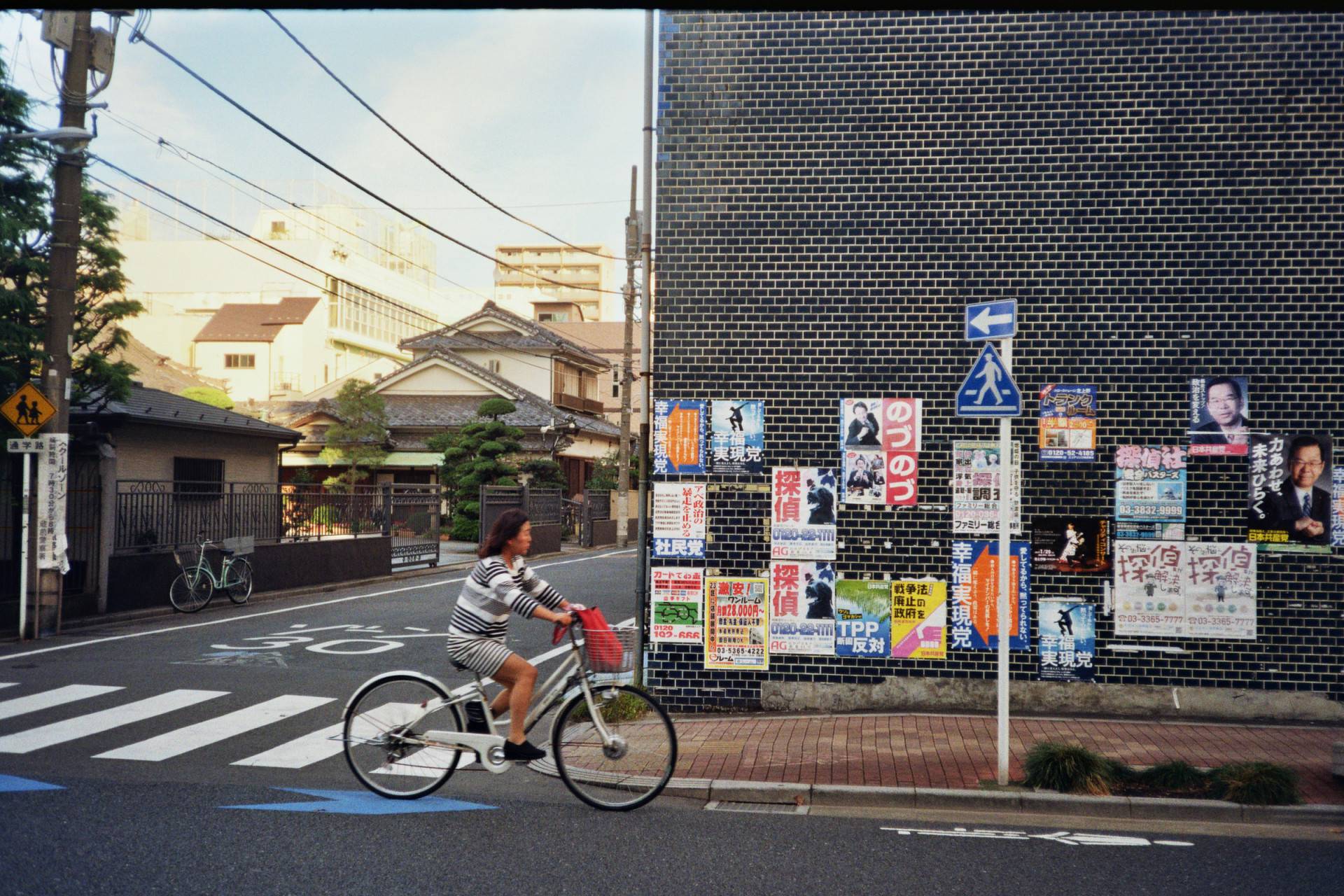 a wall on the right covered with black tiles with posters on it on the corner of a street, a woman riding a bicycle and a temple on the left side down the street