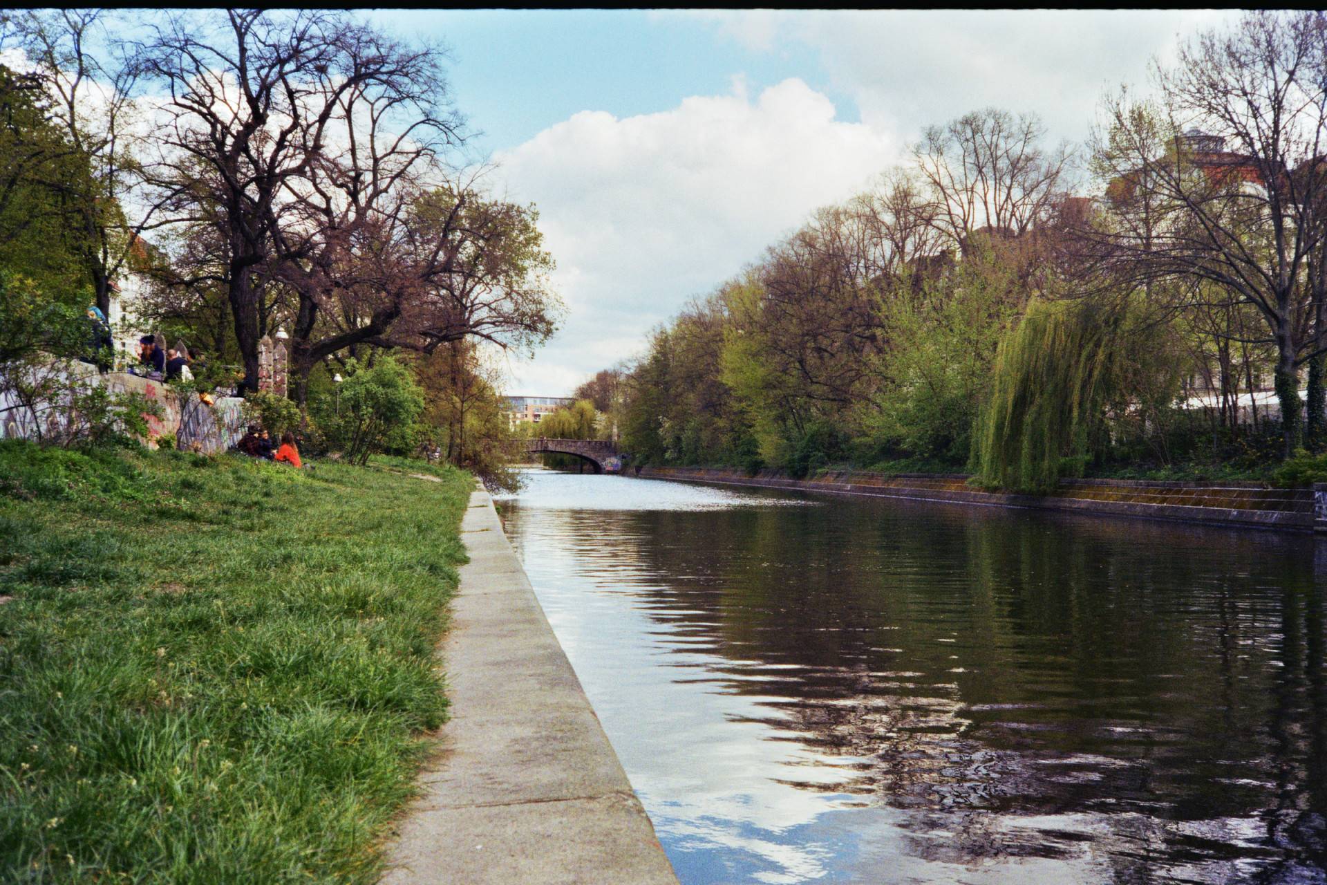 a canal surrounded with green and brown trees and grass areas, some people sitting on the left side and some buildings can be seen behind the trees