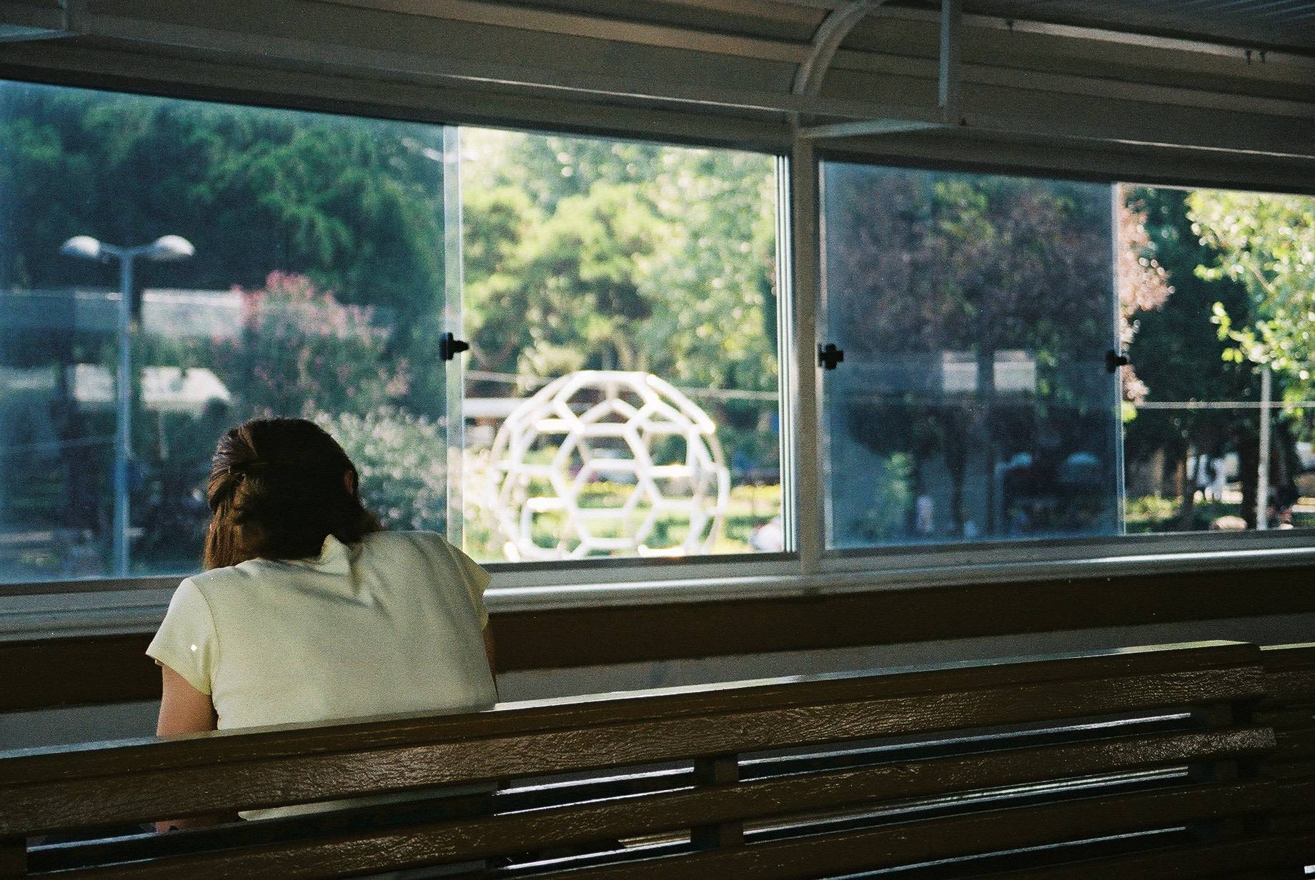 person sitting on a wooden bench seen from behind, spherical metal statue on the background seen through the window