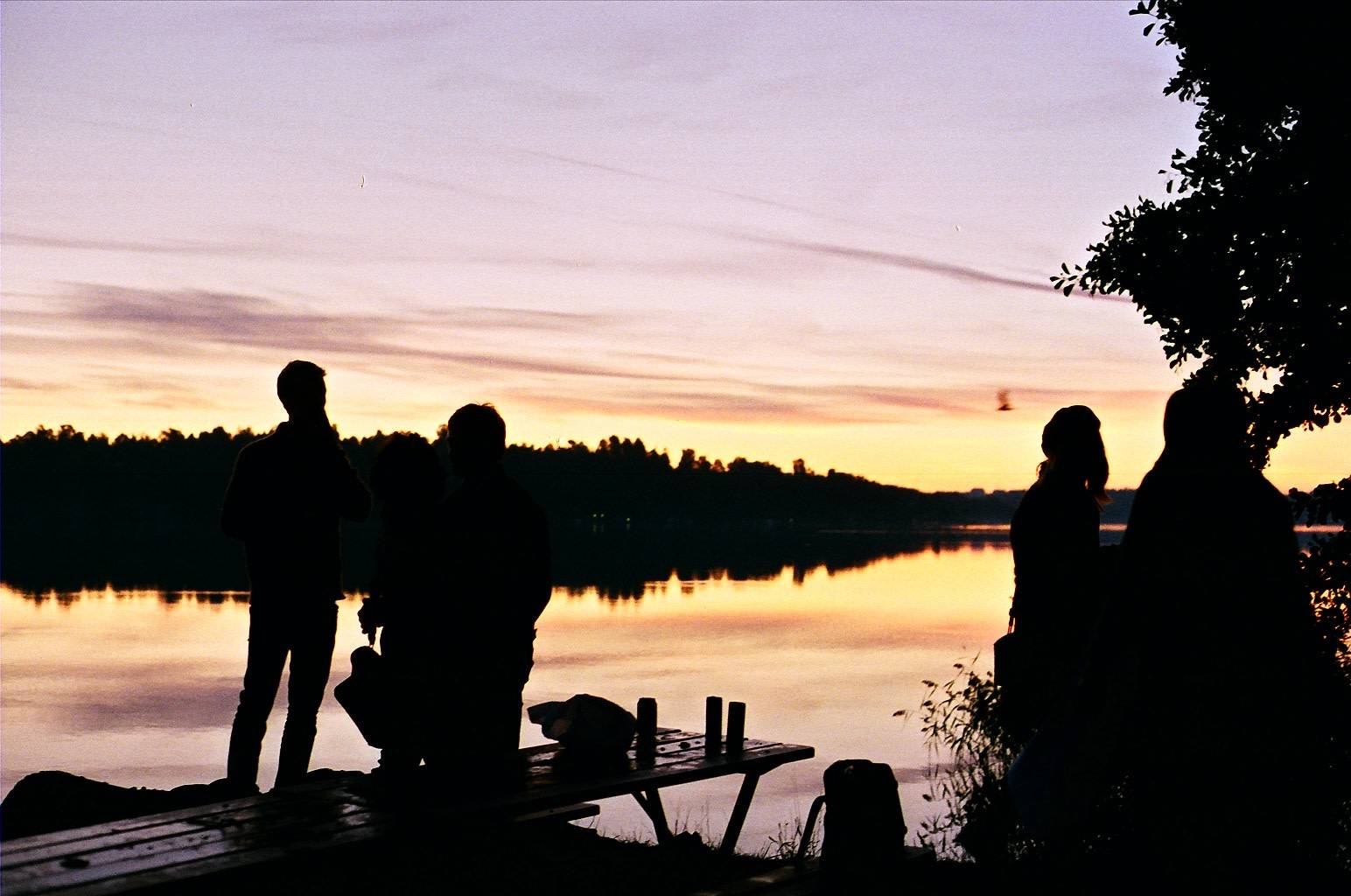 silhouettes of people hanging around the picnic table next to the lake during the sunrise