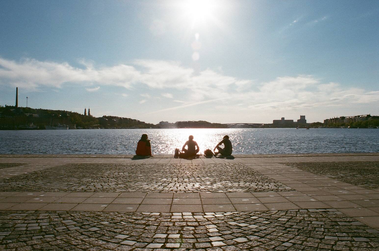 three people sitting next to the river under the sunny blue sky