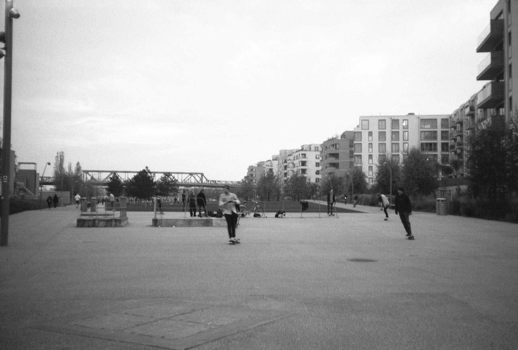 a large flat park area where several people are skateboarding, walking and hanging around, area is surrounded by buildings on the right side