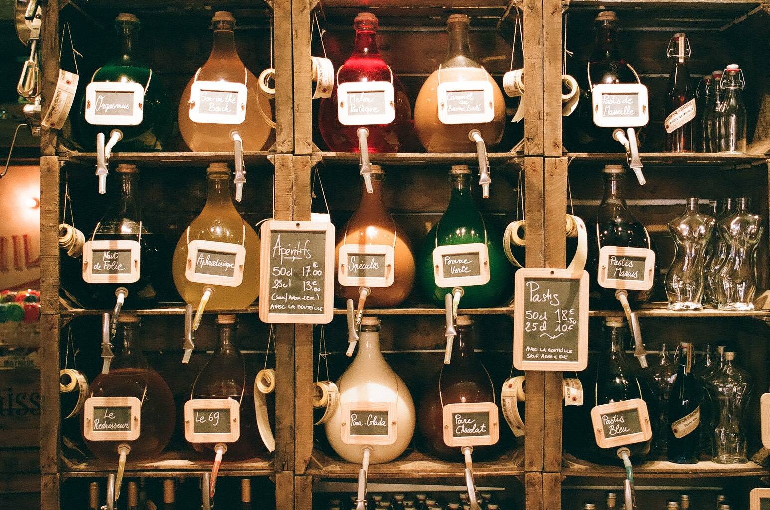 wide range of different coloured alcoholic drinks displayed in identical looking vase like glass containers that are placed on stacked wooden crates