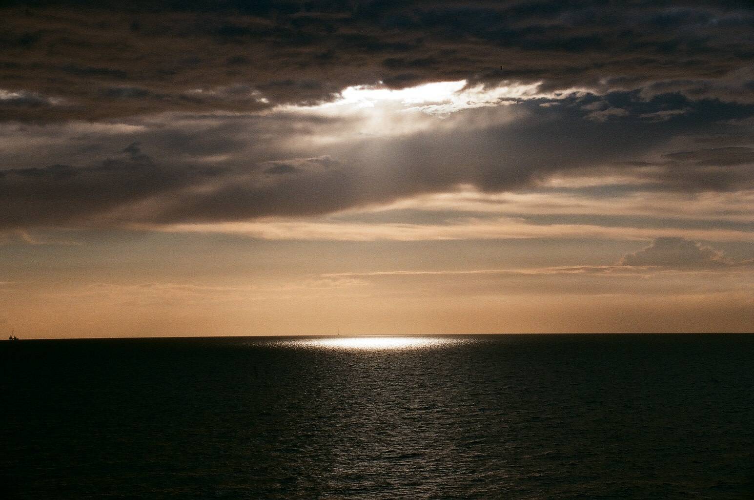 sunlight shining through the cloudy sky lightens up a small area on the dark sea, sky is clear and has orange hues on the horizon