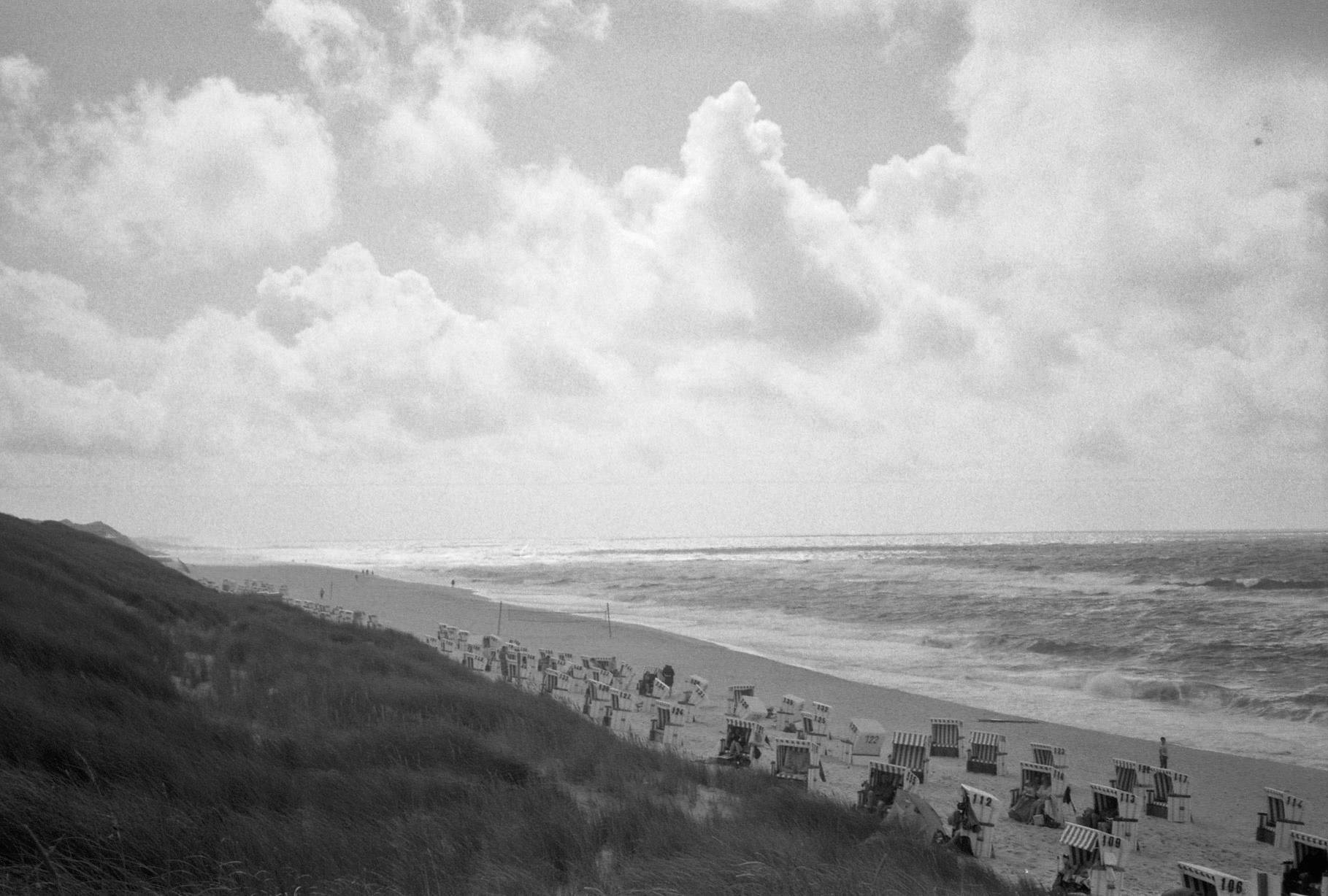 wavy see, the beach with basket chairs scattered around and the grassland behind under the bright cloudy sky