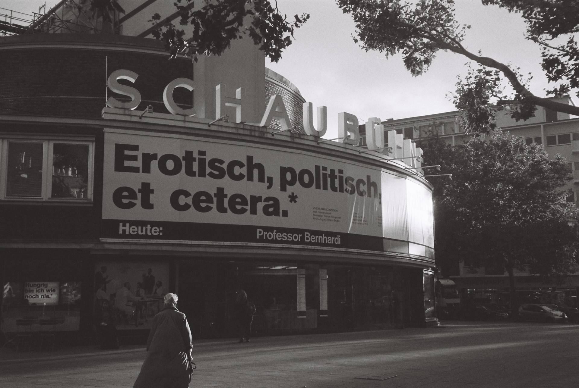 entrance of a theatre with a large sign above that reads "Erotisch, politisch, et cetera*", a person seen directly from behind is walking to the entrance