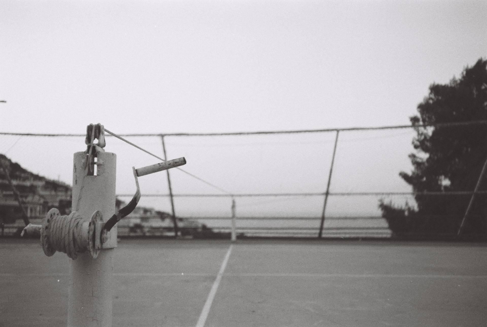 an unmaintained tennis court with the net line stretched but the net itself is missing, sea and clear sky is seen through the leaning barrier on the back