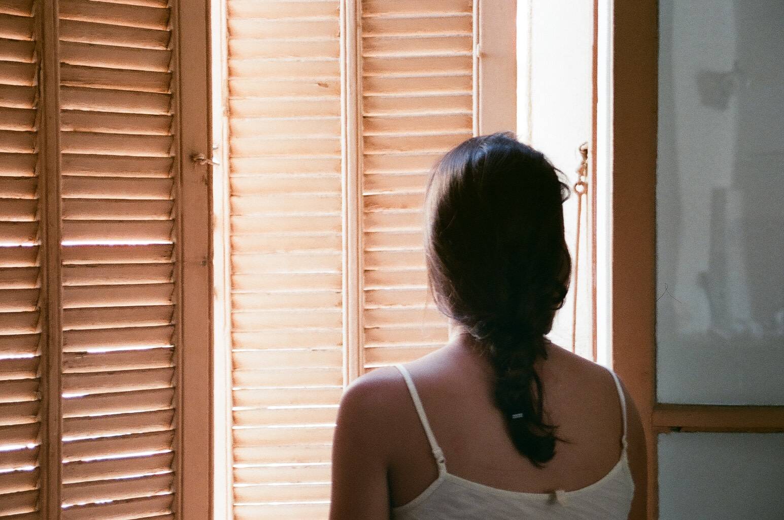 a woman seen from behind looking outside through the window where the wooden blinds are barely open